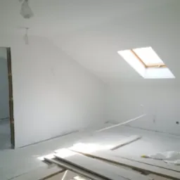 Poddasze po malowaniu na biało, widoczne deski podłogowe, okno dachowe i wystające przewody elektryczne.