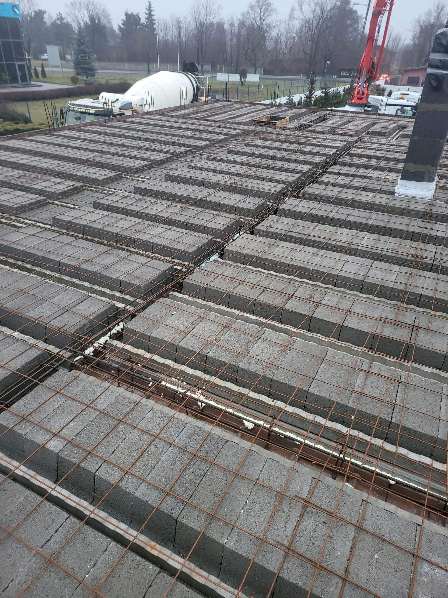 Widok z góry na strop Teriva w trakcie budowy, z widocznymi betonowymi pustakami, zbrojeniem i pianką montażową, na tle betonomieszarki i żurawia.
