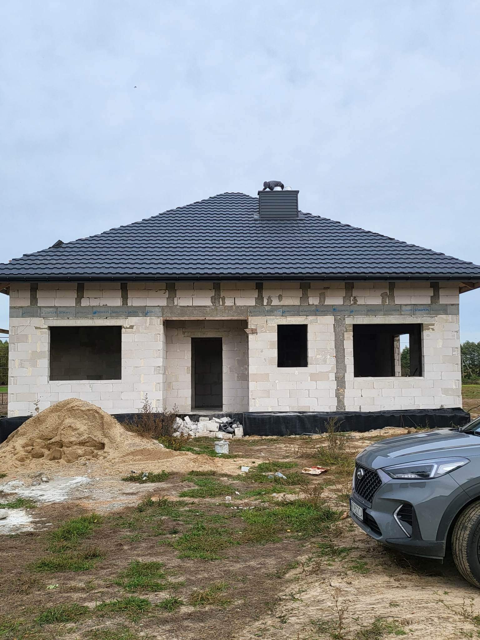 Surowy dom jednorodzinny z szarej cegły z ciemnoszarym dachem i kominem z figurką, widoczny stos piasku i fragment samochodu, budowa w toku.