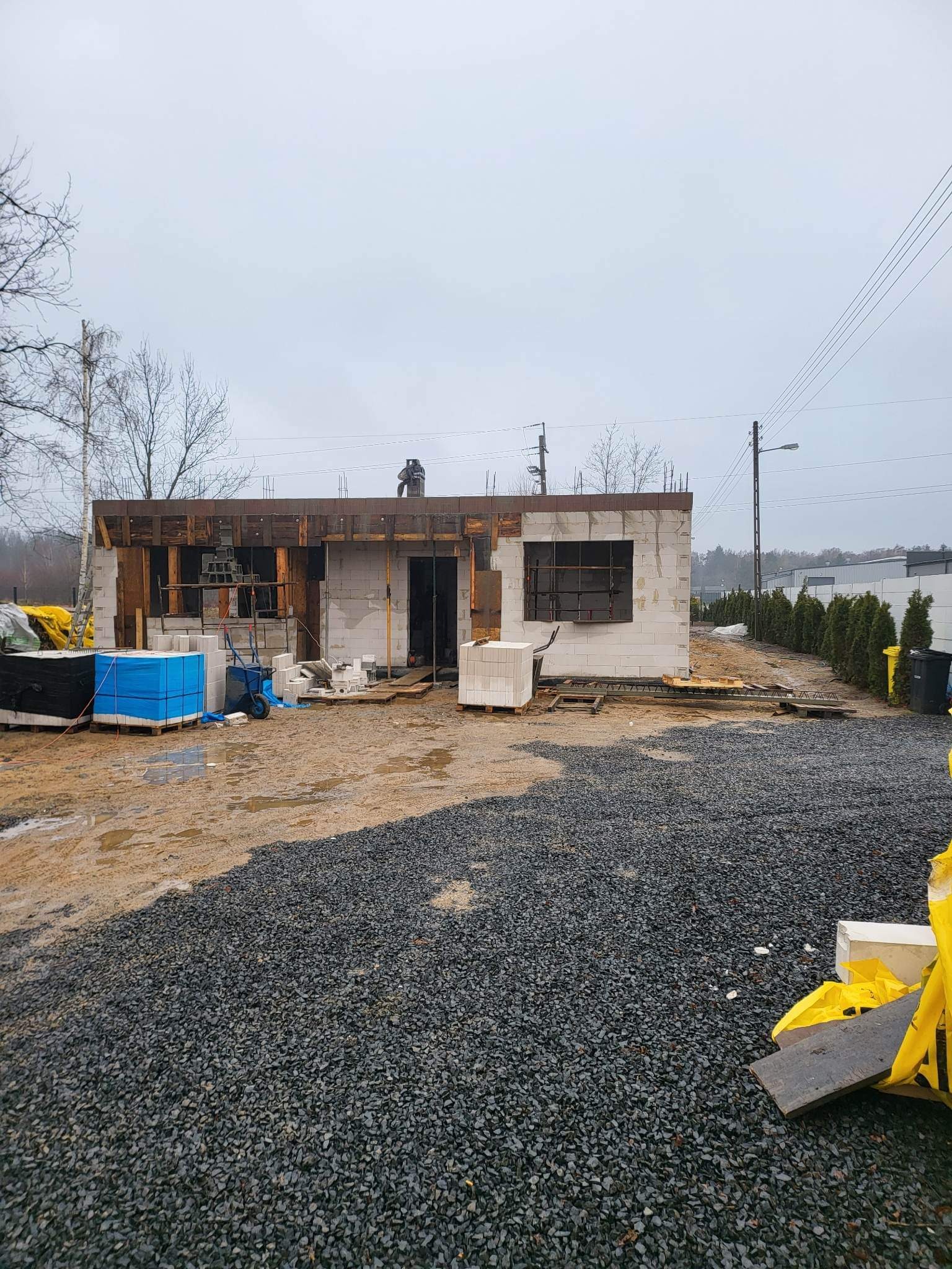 Surowy stan budowy domu z bloczków, widoczne zbrojenia i drewniana konstrukcja dachu, teren wokół budynku z gruzem i materiałami budowlanymi, pochmurny dzień.