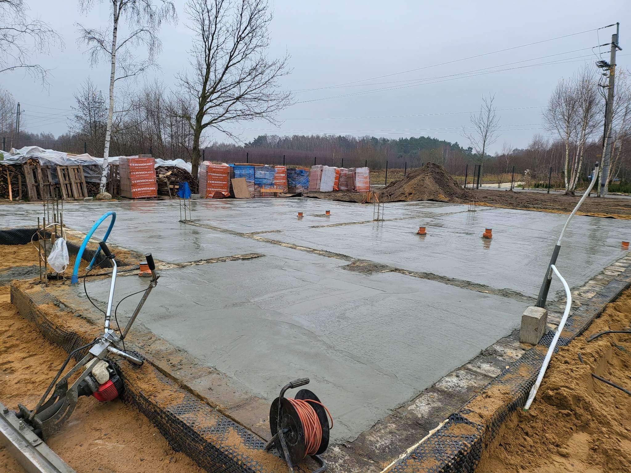 Wylany fundament pod dom jednorodzinny z widocznym zagęszczarką powierzchniową i stertą cegieł w tle, pochmurny dzień.
