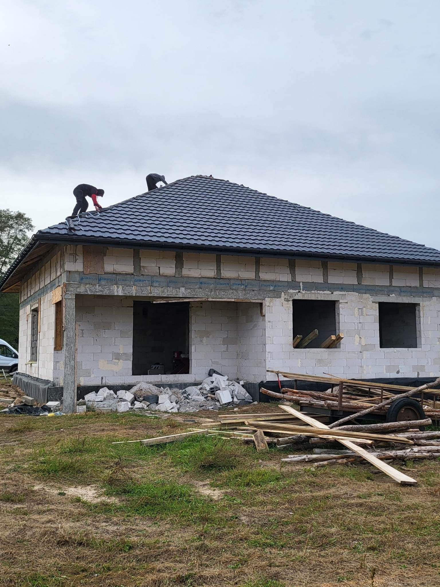 Wznoszenie domu z białych bloczków: dwóch robotników montuje ciemnoszarą dachówkę na nowym dachu, widoczne niezabezpieczone otwory okienne i drzwiowe, porozrzucane materiały budowlane na trawie.