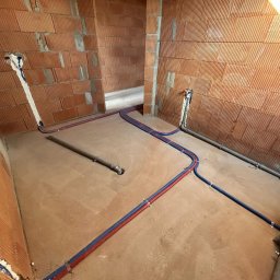 Karat Instal Krzysztof Czyż - Wnętrze w trakcie budowy z instalacją hydrauliczną: rury czerwono-niebieskie ułożone na podłodze, pionowe podejścia do ściany z cegły dziurawki. Widoczne mocowania rur.