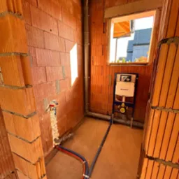 Surowy stan łazienki w trakcie instalacji: widoczne rury, stelaż WC Geberit na tle ceglanych ścian i wylewki. Instalacja wodno-kanalizacyjna w nowym budynku.