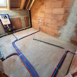 Karat Instal Krzysztof Czyż - Instalacja wodna w surowym wnętrzu z czerwoną cegłą i oknem dachowym. Widoczne rury, stelaż podtynkowy Geberit i fragment instalacji kanalizacyjnej.