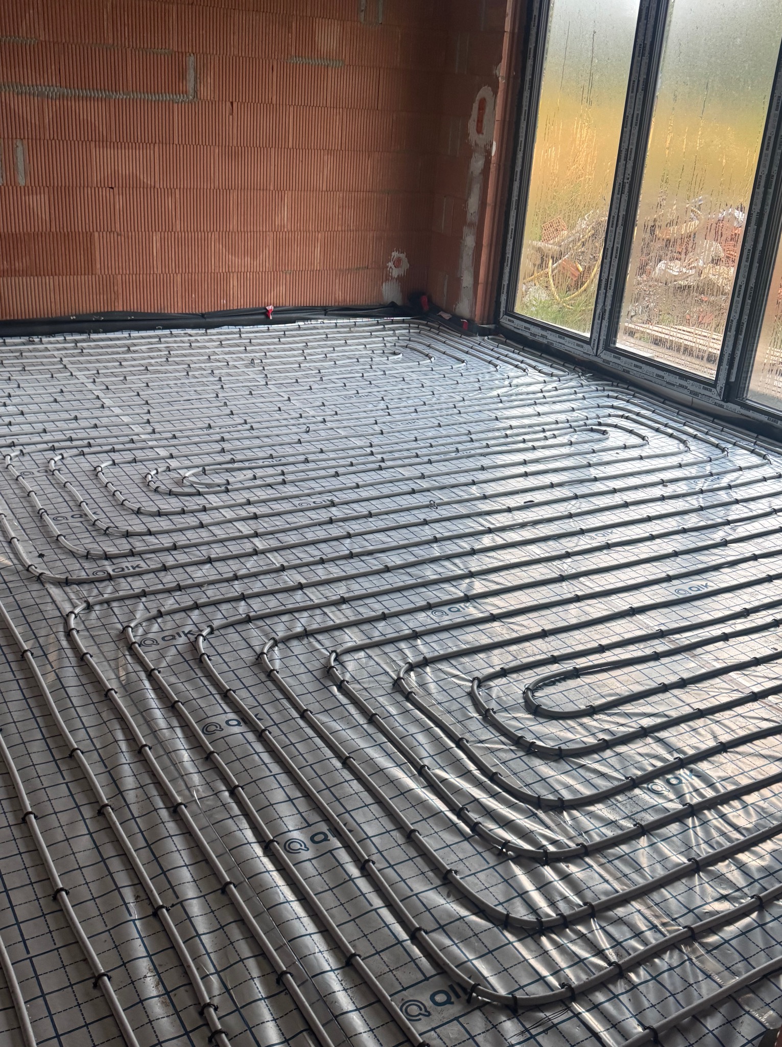 Szare rury ogrzewania podłogowego ułożone na folii z nadrukiem kratki, widok z góry, ściany z cegły i okno w tle.