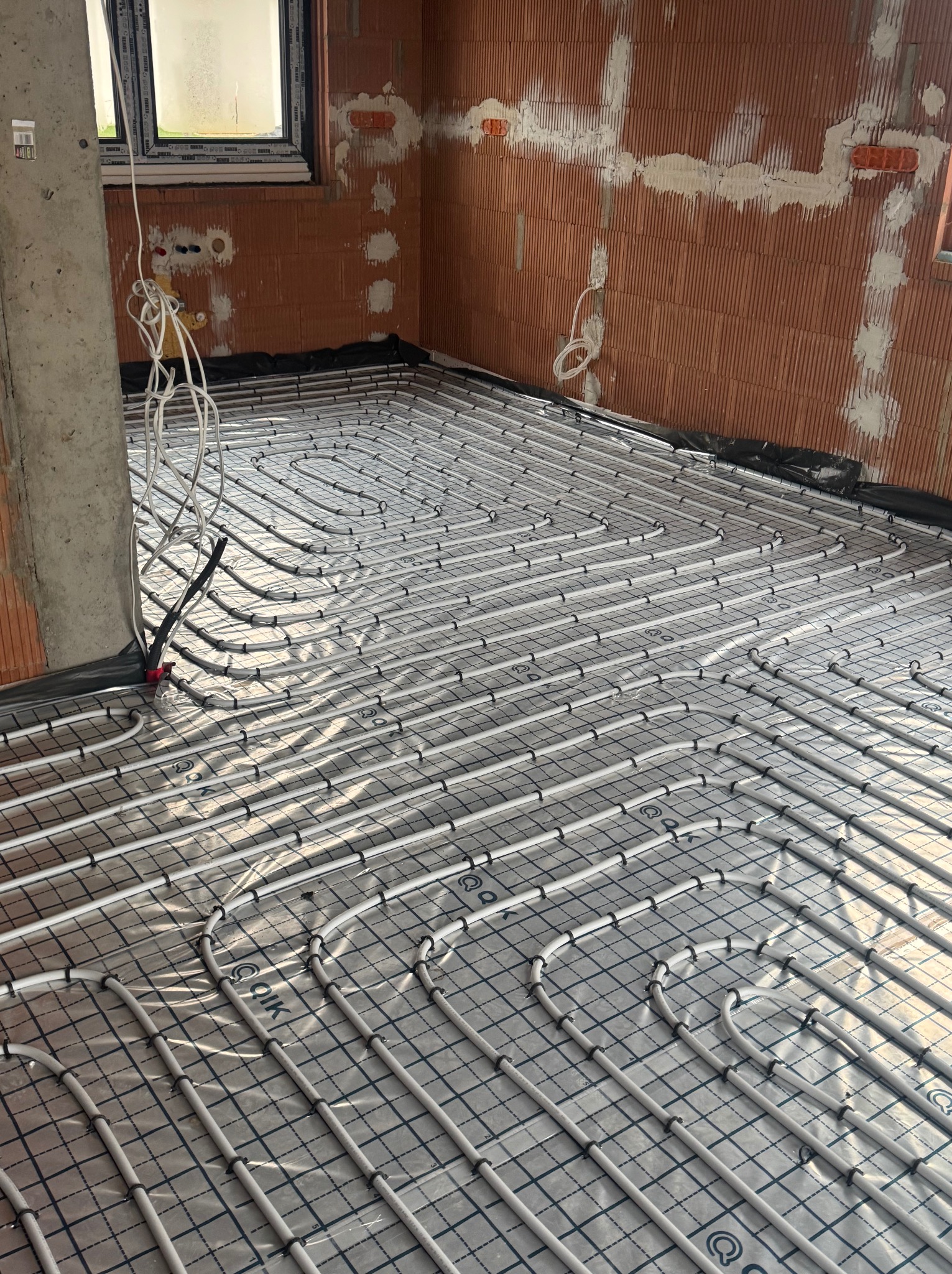 Rozłożone rury ogrzewania podłogowego na folii izolacyjnej w surowym pomieszczeniu z ceglanymi ścianami. Widoczne mocowania rur i wystające przewody.