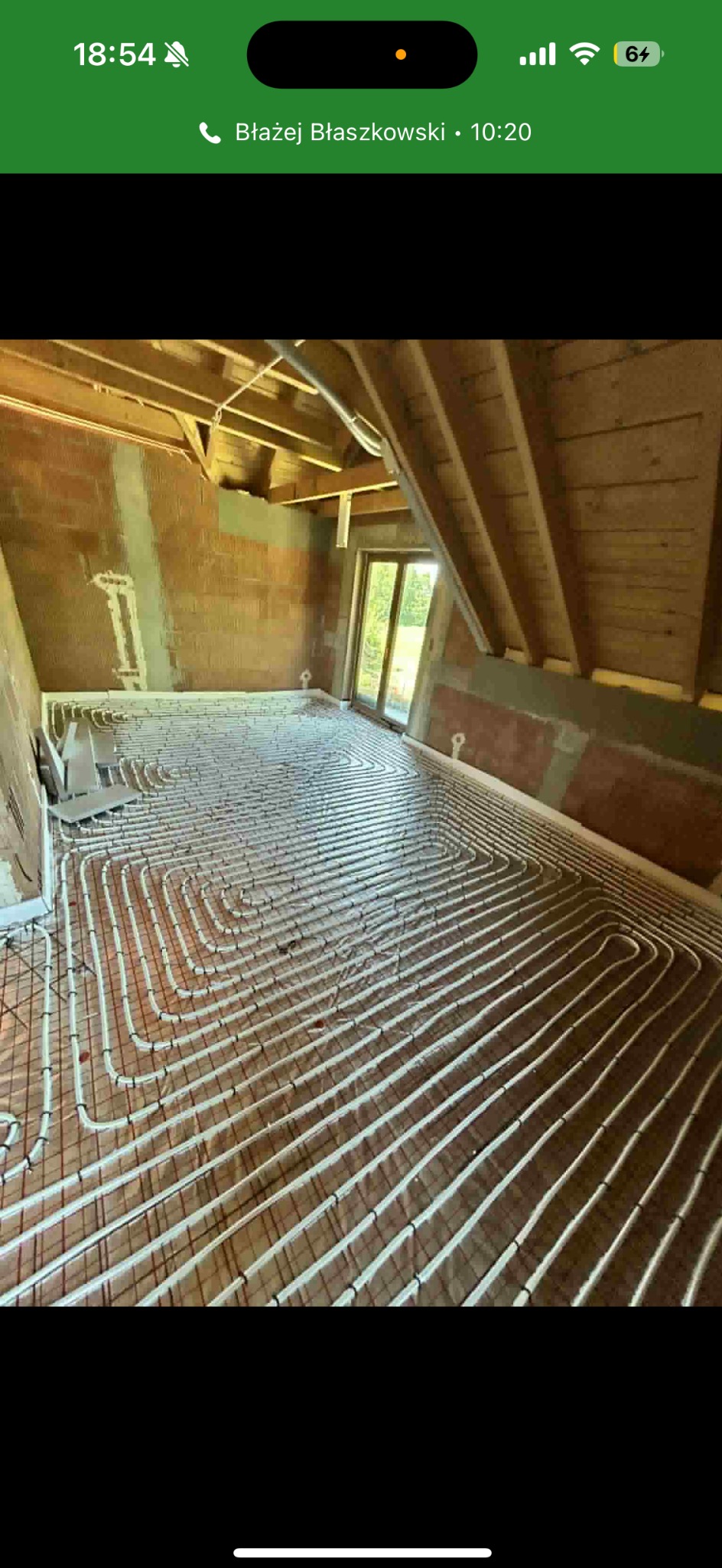 Podłogówka w surowym wnętrzu poddasza: białe rury ułożone w pętle na folii, widoczne ceglane ściany i drewniana konstrukcja dachu, okno z widokiem na zieleń.