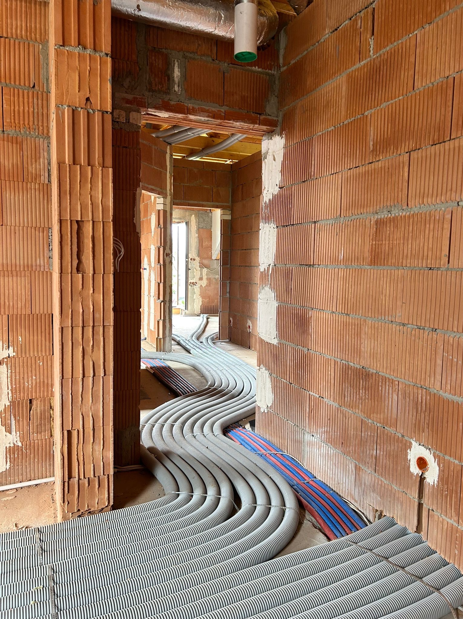 Instalacja ogrzewania podłogowego w surowym wnętrzu. Rury grzewcze ułożone na izolacji, widoczne ściany z czerwonej cegły i otwory drzwiowe. Etap prac przed wylewką.