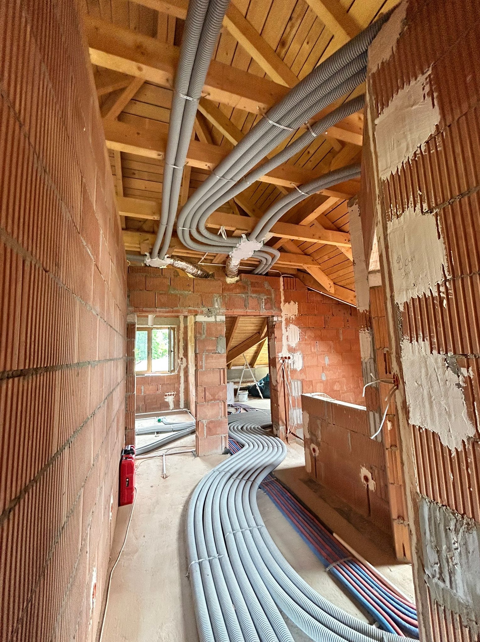 Wnętrze w trakcie budowy z widoczną instalacją ogrzewania podłogowego i instalacjami pod stropem. Rury w peszlach prowadzone wzdłuż ścian z cegły.