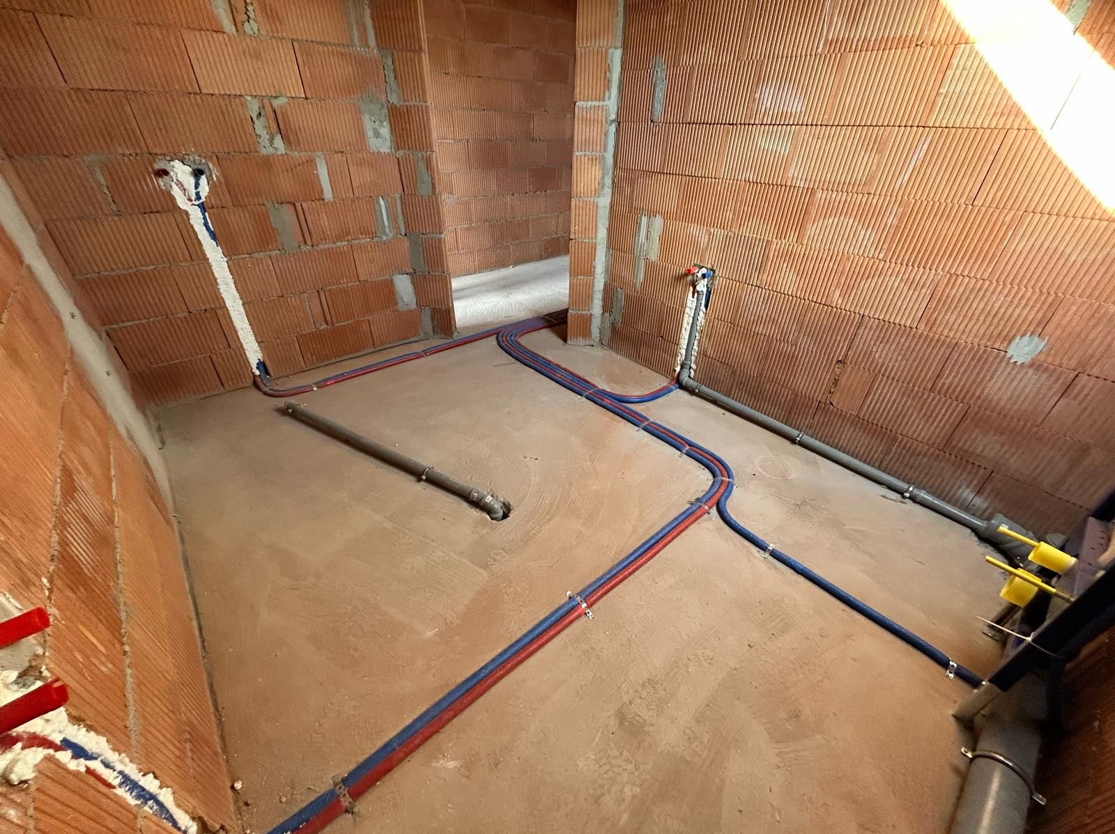 Wnętrze w trakcie budowy z instalacją hydrauliczną: rury czerwono-niebieskie ułożone na podłodze, pionowe podejścia do ściany z cegły dziurawki. Widoczne mocowania rur.