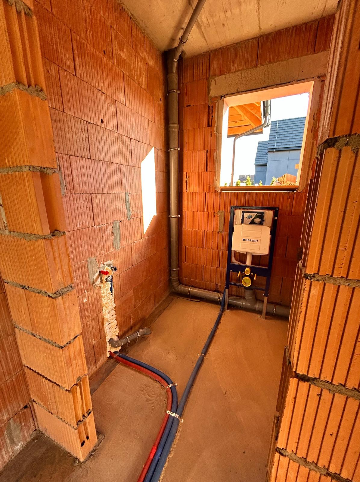 Surowy stan łazienki w trakcie instalacji: widoczne rury, stelaż WC Geberit na tle ceglanych ścian i wylewki. Instalacja wodno-kanalizacyjna w nowym budynku.