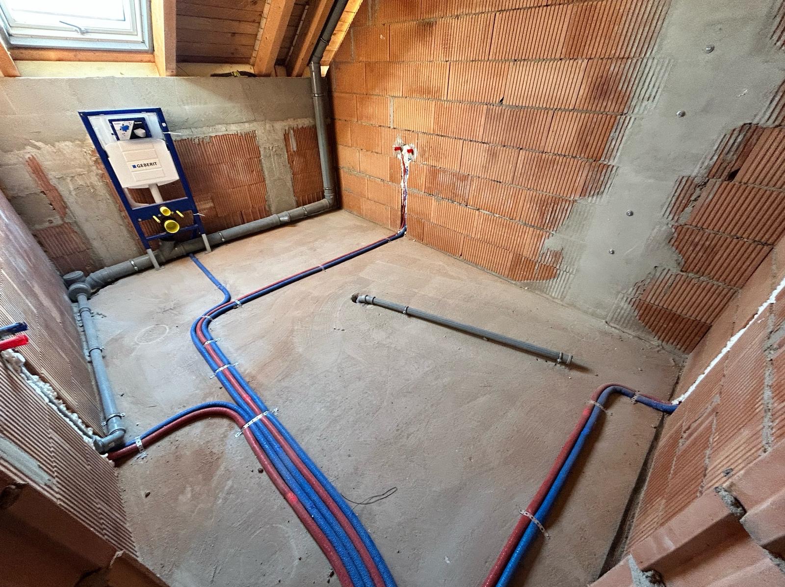 Instalacja wodna w surowym wnętrzu z czerwoną cegłą i oknem dachowym. Widoczne rury, stelaż podtynkowy Geberit i fragment instalacji kanalizacyjnej.