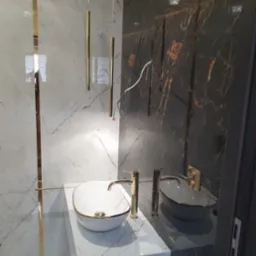 Elegancka łazienka z dwoma umywalkami nablatowymi: białą z złotym obramowaniem i szarą, na marmurowych blatach, z złotymi bateriami i wiszącymi lampami tubami na tle marmurowych ścian w odcieniach...