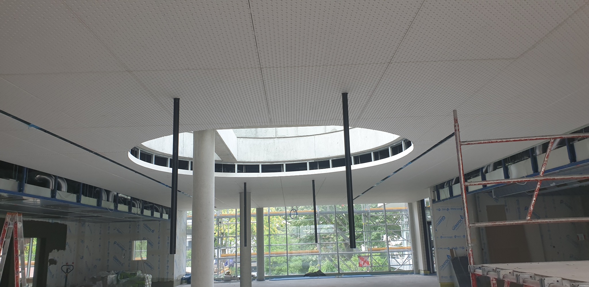 Montaż sufitu podwieszanego z okrągłym świetlikiem w budynku komercyjnym, widoczne płyty z perforacją, czarne elementy konstrukcyjne i rusztowanie.