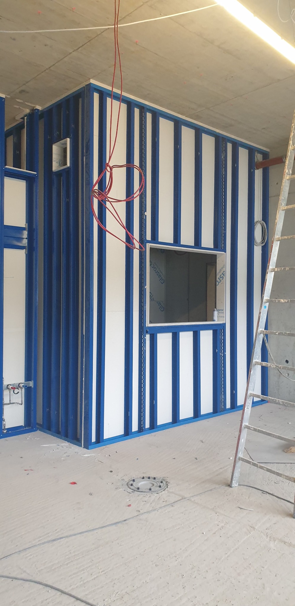 Wnętrze w trakcie prac wykończeniowych: konstrukcja ścian z niebieskich profili i białych paneli, okno w ścianie, widoczne kable elektryczne i drabina.