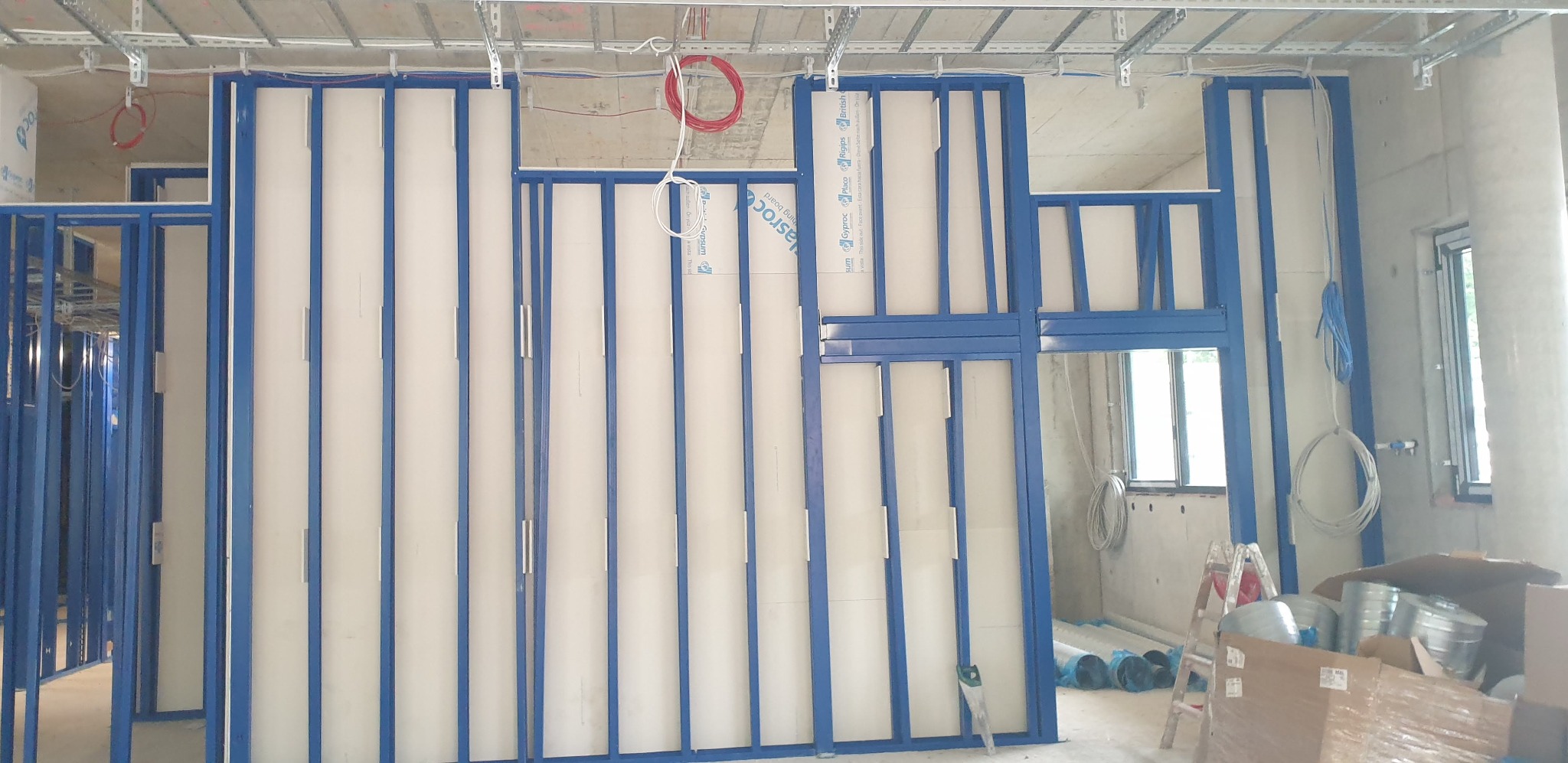 Konstrukcja szkieletowa z profili stalowych w kolorze niebieskim, wypełniona białymi płytami gipsowo-kartonowymi, widoczne przewody elektryczne i sufit podwieszany, w tle okno i materiały budowlane.