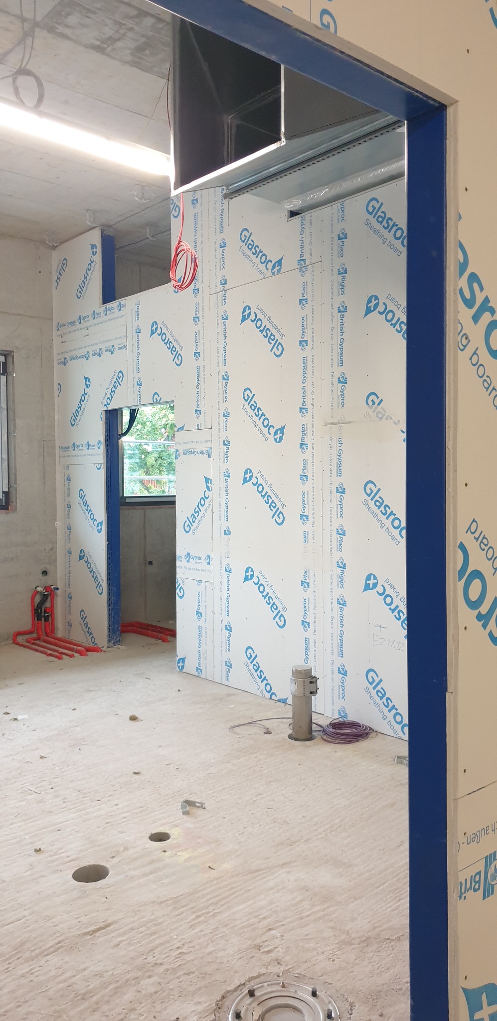 Wnętrze w trakcie prac wykończeniowych: ściany obłożone płytami Glasroc, widoczne instalacje elektryczne i hydrauliczne, surowa podłoga z otworami.