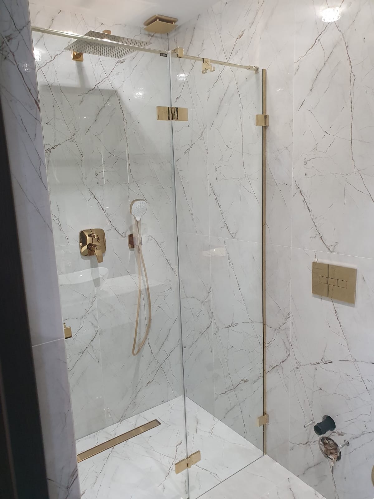 Kabina prysznicowa z białymi marmurowymi płytkami, złotymi okuciami i deszczownicą, widoczne elementy armatury i odpływ liniowy.