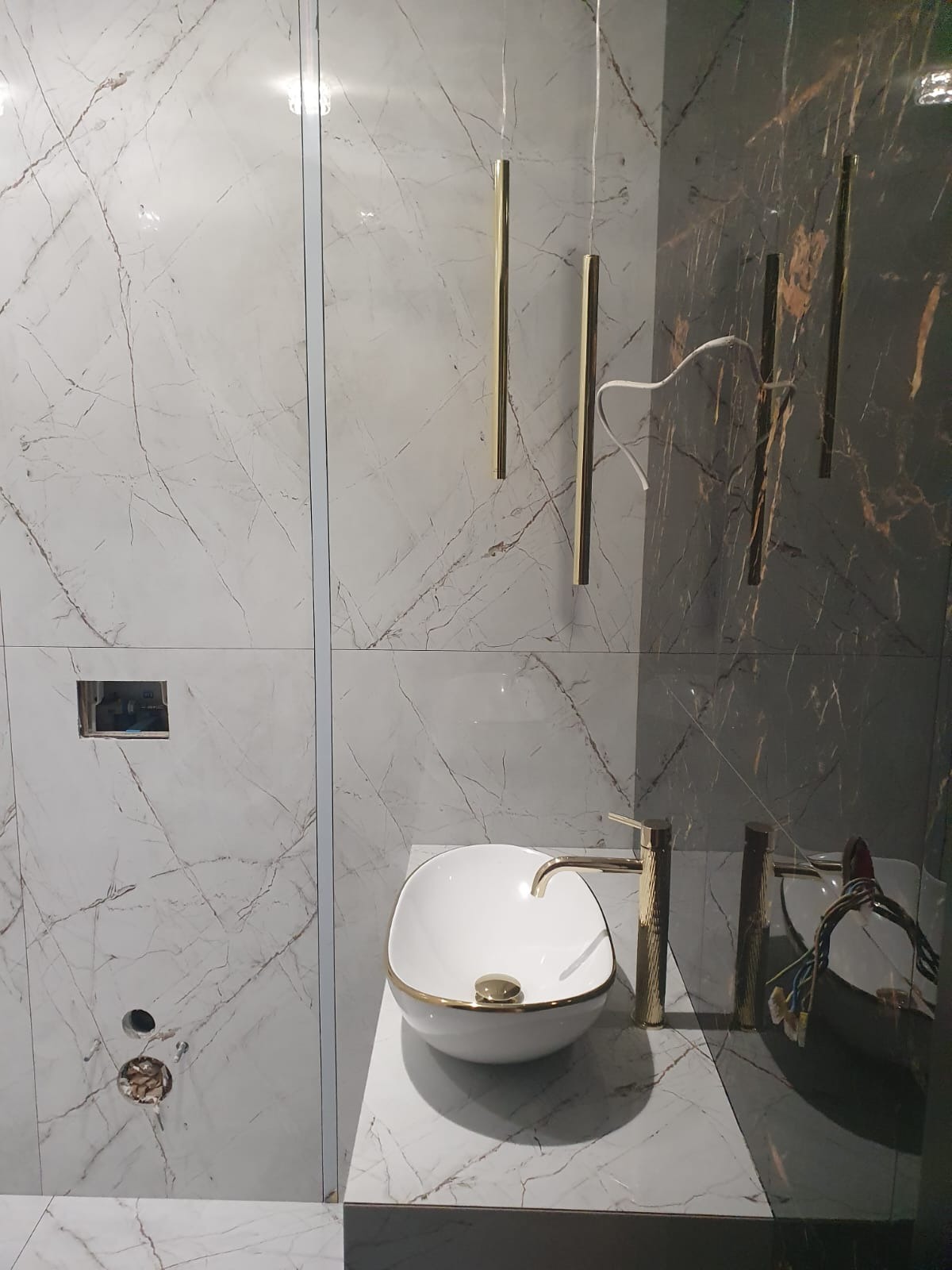 Montaż umywalki nablatowej z baterią w łazience wykończonej płytkami imitującymi marmur, widoczne otwory na przyłącza i elementy oświetlenia.