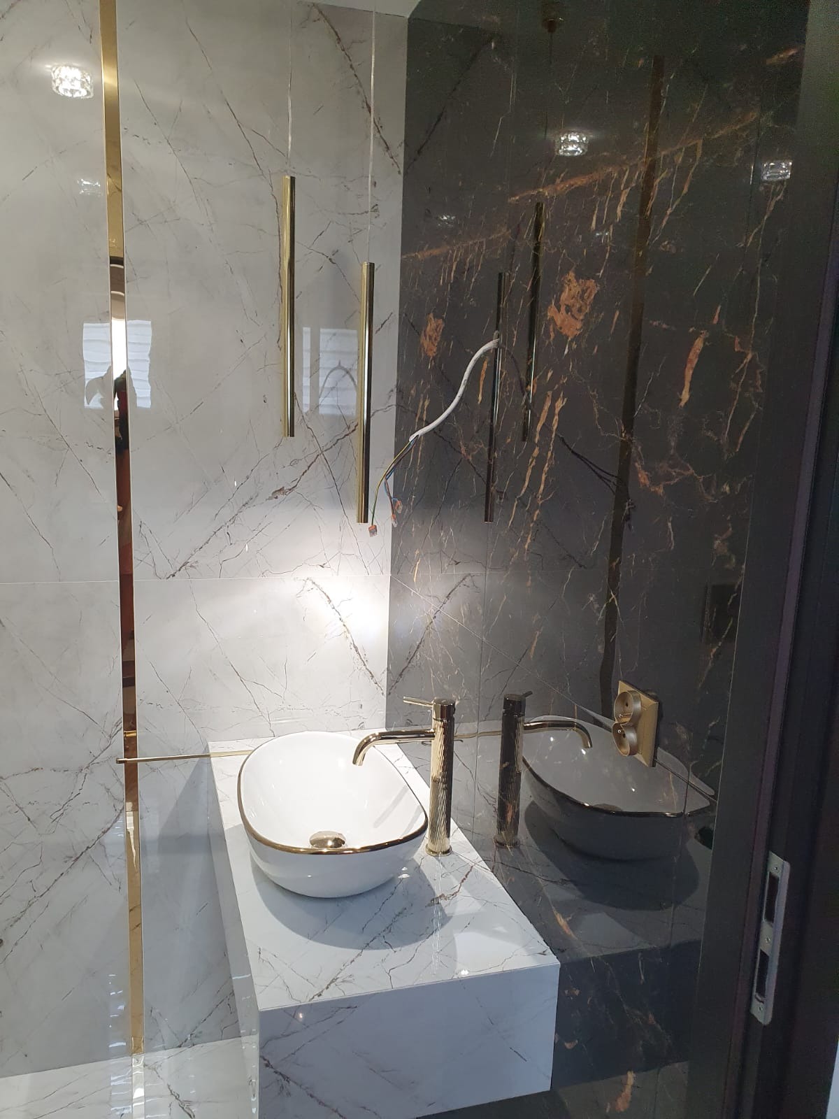 Elegancka łazienka z dwoma umywalkami nablatowymi: białą z złotym obramowaniem i szarą, na marmurowych blatach, z złotymi bateriami i wiszącymi lampami tubami na tle marmurowych ścian w odcieniach...