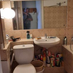 M-Tech Maksym Dykunets - Umywalka, toaleta i lustro w łazience z beżowymi płytkami. W lustrze odbija się osoba robiąca zdjęcie. Pod umywalką widoczne środki czystości.