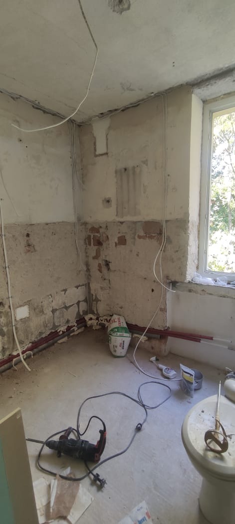 Wnętrze w trakcie remontu: odsłonięta cegła, instalacje, worki z zaprawą, narzędzia (wiertarka, mieszadło). Okno z widokiem na zieleń. Ściany częściowo przygotowane do gładzi.