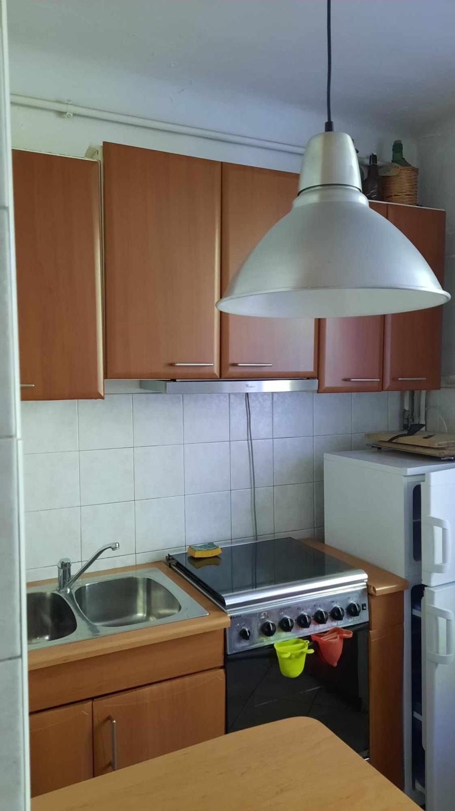 Kuchnia z brązowymi szafkami, białą lodówką i dużym, metalowym kloszem lampy. Widoczny zlew, płyta grzewcza i białe płytki na ścianie.