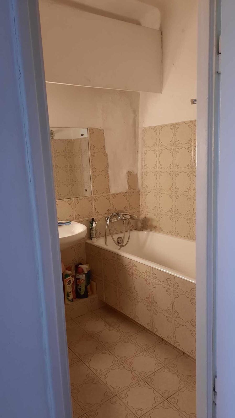 Beżowa łazienka z wanną w płytkach, umywalką i lustrem. Widoczne środki czystości. Ujęcie przez otwarte drzwi, z fragmentem futryny po obu stronach.