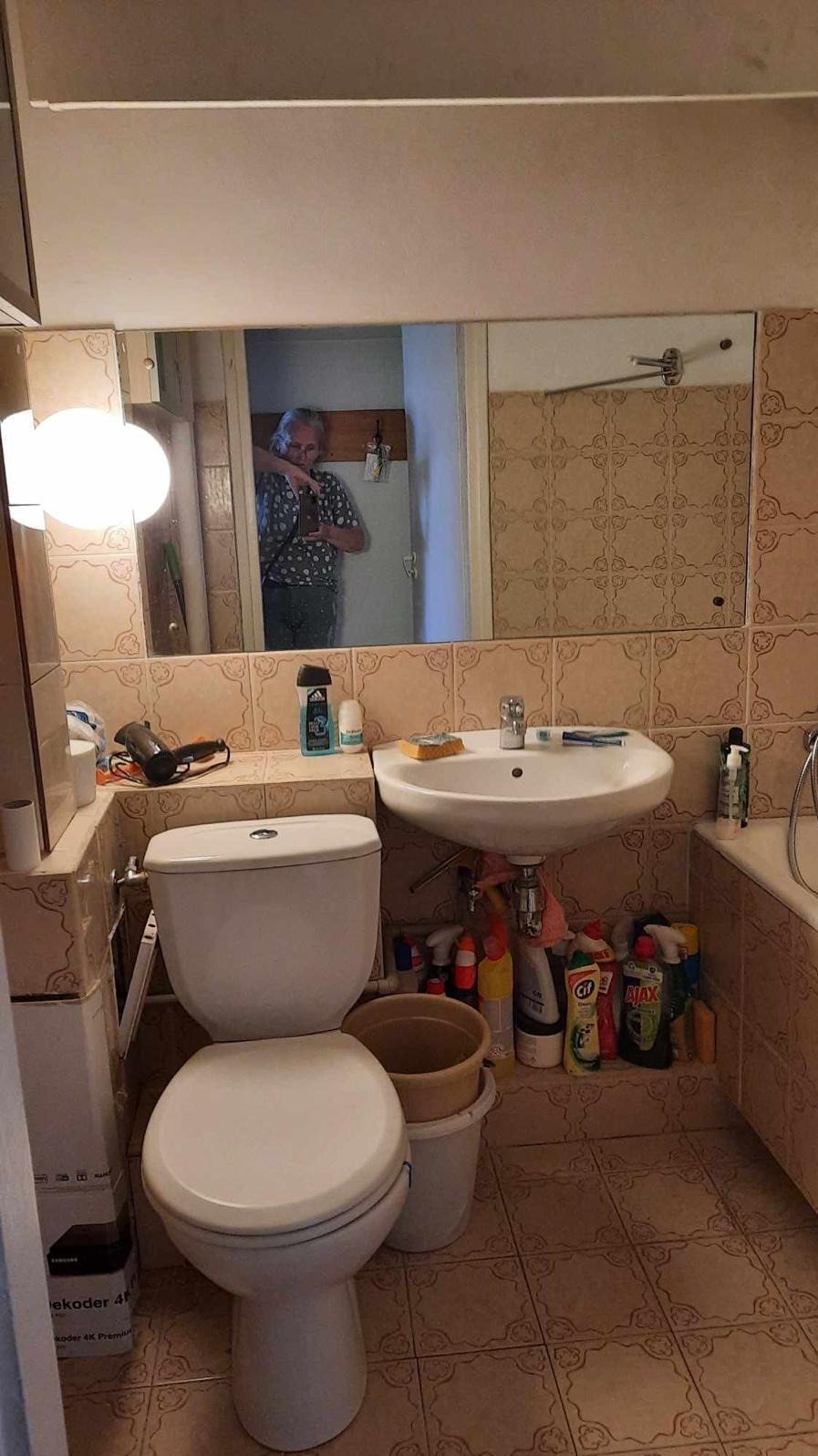 Umywalka, toaleta i lustro w łazience z beżowymi płytkami. W lustrze odbija się osoba robiąca zdjęcie. Pod umywalką widoczne środki czystości.