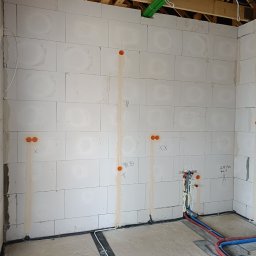 KD Install - Surowy stan wnętrza z instalacjami: widoczne przewody elektryczne w ścianie z betonu komórkowego, rury wodne w podłodze i pod sufitem, zielona rura karbowana pod stropem drewnianym.