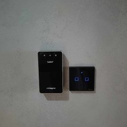 KD Install - Czarny panel sterowania Integra Safet z ikonami alarmu, oka i dzwonka oraz czarny dotykowy włącznik światła Sonoff z niebieskimi podświetlanymi ikonami, zamontowane na szarej ścianie o chropowatej...