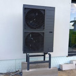 KD Install - Zewnętrzna jednostka pompy ciepła Panasonic Aquarea zamontowana na wsporniku przy białej ścianie budynku, widoczne elementy instalacji i otoczenie budowy.