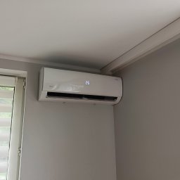 KD Install - Biały klimatyzator Rotenso z wyświetlaczem 26°C zamontowany na szarej ścianie w pokoju, widok z dołu. Obok okno z żaluzjami dzień-noc.