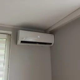 Biały klimatyzator Rotenso z wyświetlaczem 26°C zamontowany na szarej ścianie w pokoju, widok z dołu. Obok okno z żaluzjami dzień-noc.