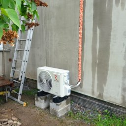 KD Install - Biały klimatyzator Rotenso na betonowych blokach przy ścianie budynku z pionową linią pianki montażowej, drabina oparta o ścianę, widok z zewnątrz.