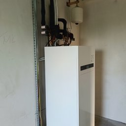 KD Install - Biała pompa ciepła z czarnymi rurami i zbiornikiem wyrównawczym na ścianie w surowym wnętrzu, widoczne przewody elektryczne w korytku.