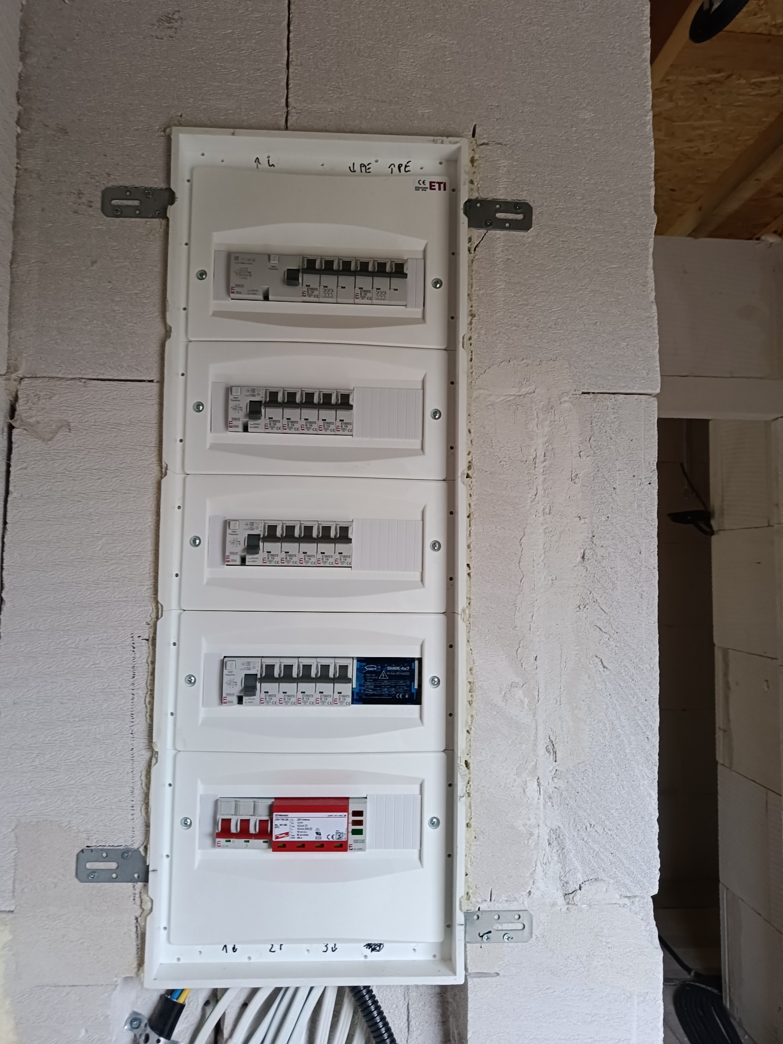 Rozdzielnica elektryczna, pięć modułów, zamontowana na ścianie z betonu komórkowego, widoczne przewody i metalowe uchwyty mocujące.