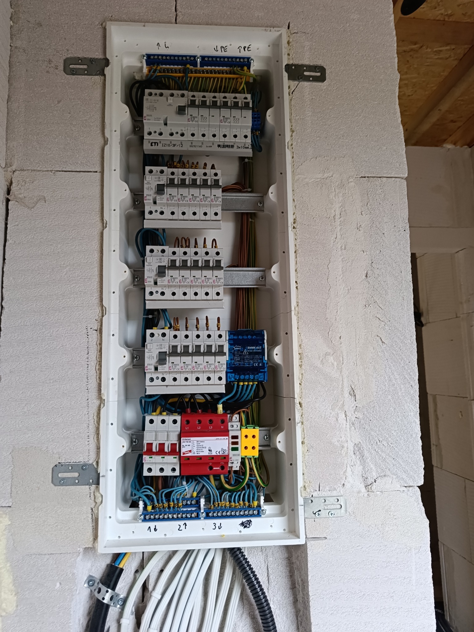 Rozdzielnica elektryczna z zabezpieczeniami zamontowana na ścianie z betonu komórkowego, widoczne przewody i aparatura modułowa.