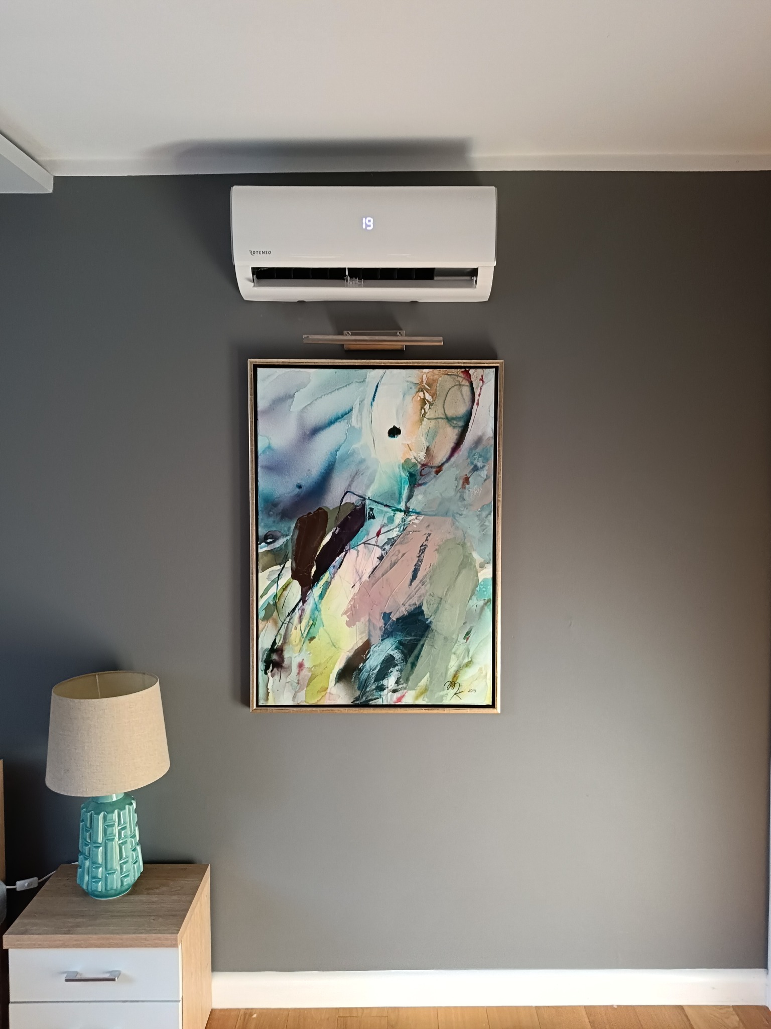 Biała klimatyzacja ścienna, ustawiona nad abstrakcyjnym obrazem w złotych ramach, oświetlona lampką; na szafce nocnej w Świebodzicach stoi turkusowa lampka. Widoczna temperatura 19 stopni.