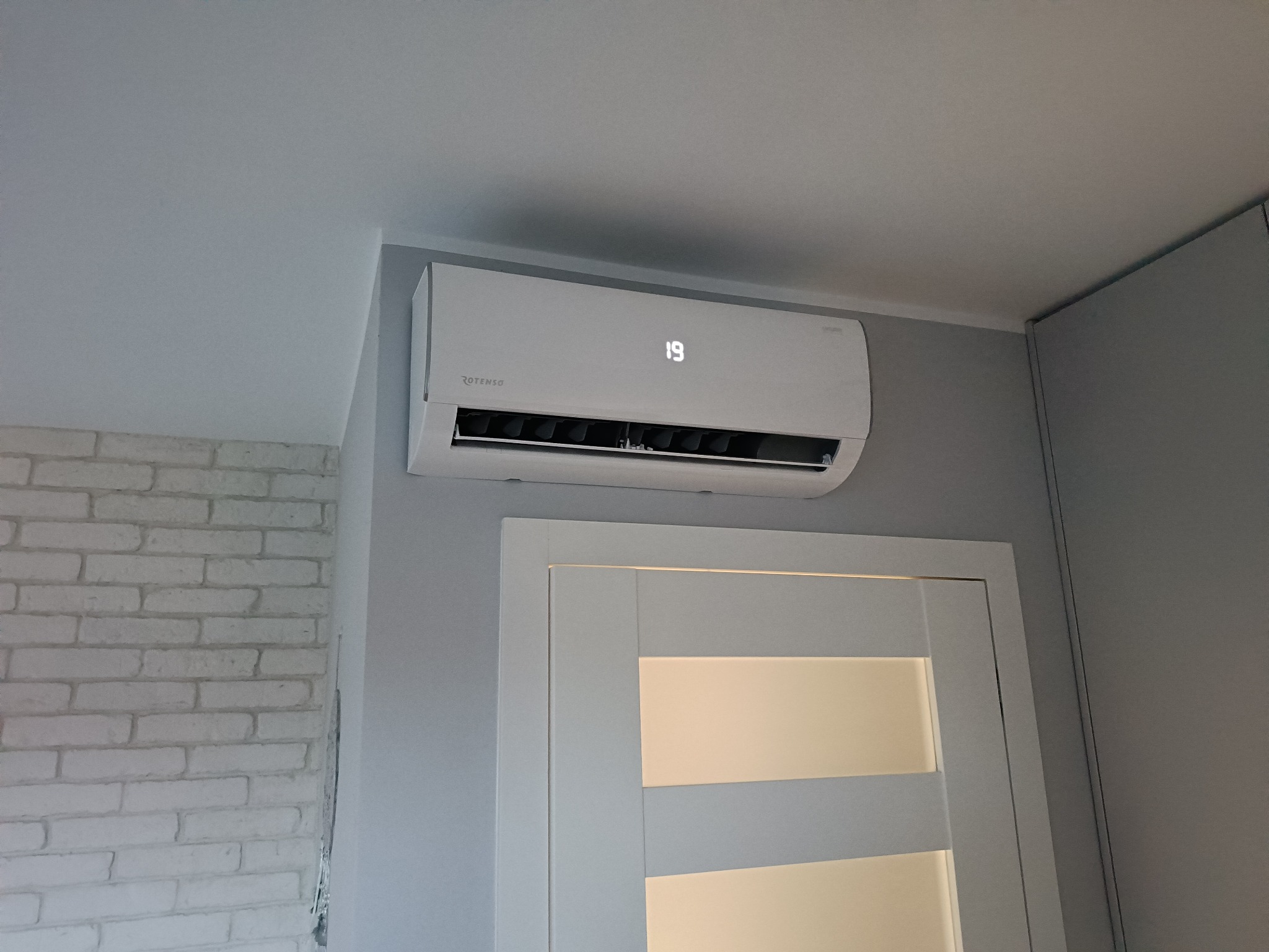 Biały klimatyzator Rotenso z wyświetlaczem temperatury 19 stopni Celsjusza, zamontowany na szarej ścianie nad białymi drzwiami wewnętrznymi z poziomymi panelami, obok ściany z białej cegły.