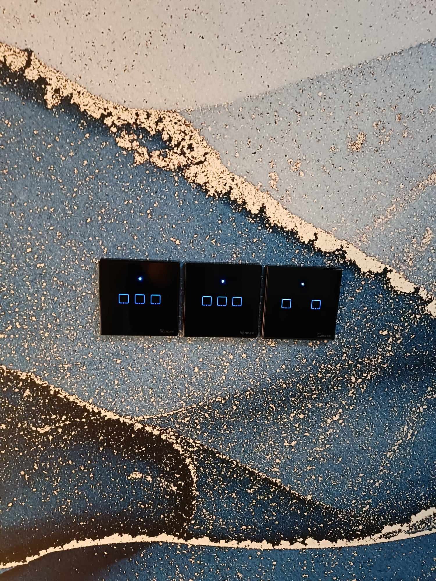 Trzy czarne, dotykowe włączniki światła Sonoff z niebieskimi diodami LED, zamontowane na ścianie z abstrakcyjnym, niebiesko-białym wzorem.
