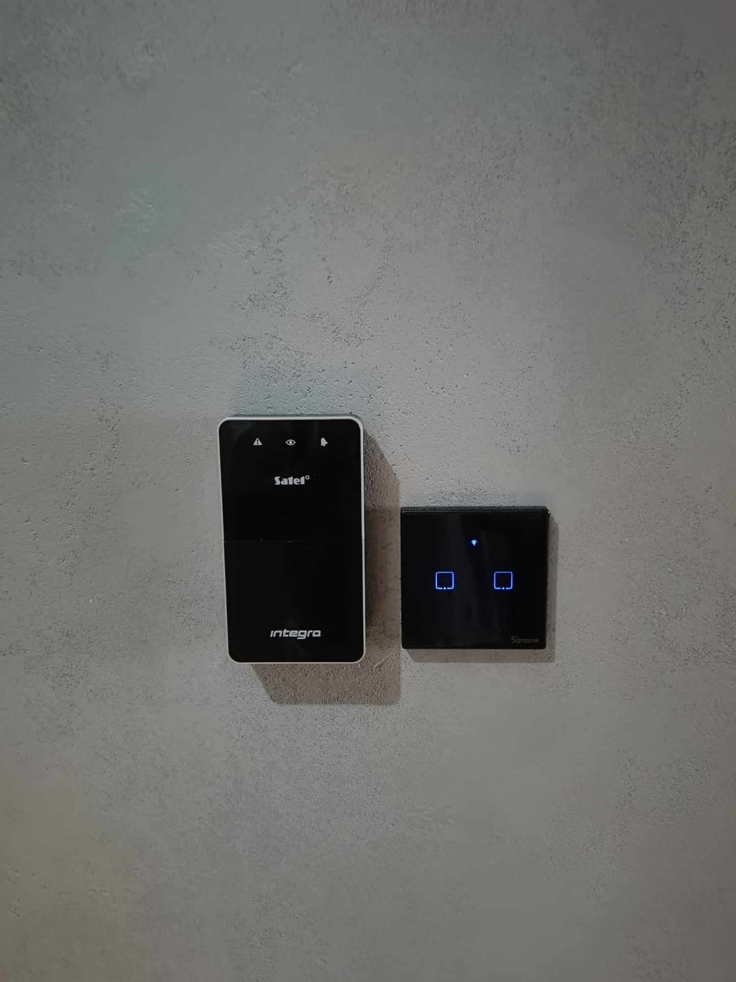 Czarny panel sterowania Integra Safet z ikonami alarmu, oka i dzwonka oraz czarny dotykowy włącznik światła Sonoff z niebieskimi podświetlanymi ikonami, zamontowane na szarej ścianie o chropowatej...