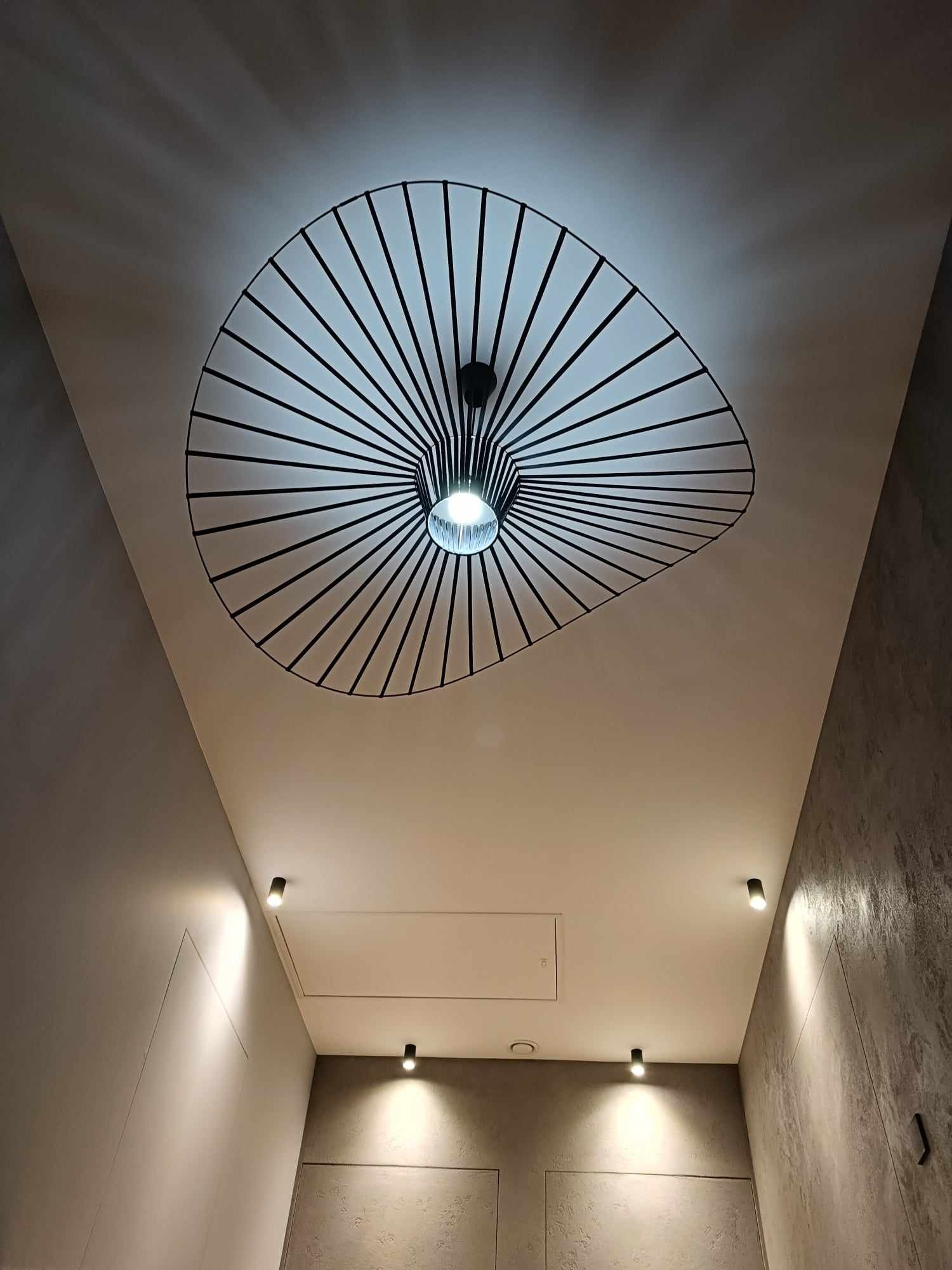Nowoczesna lampa sufitowa z czarnymi prętami tworzącymi okrągły wzór, uzupełniona o punktowe oświetlenie w aranżacji wnętrza z szarą ścianą o fakturze betonu.