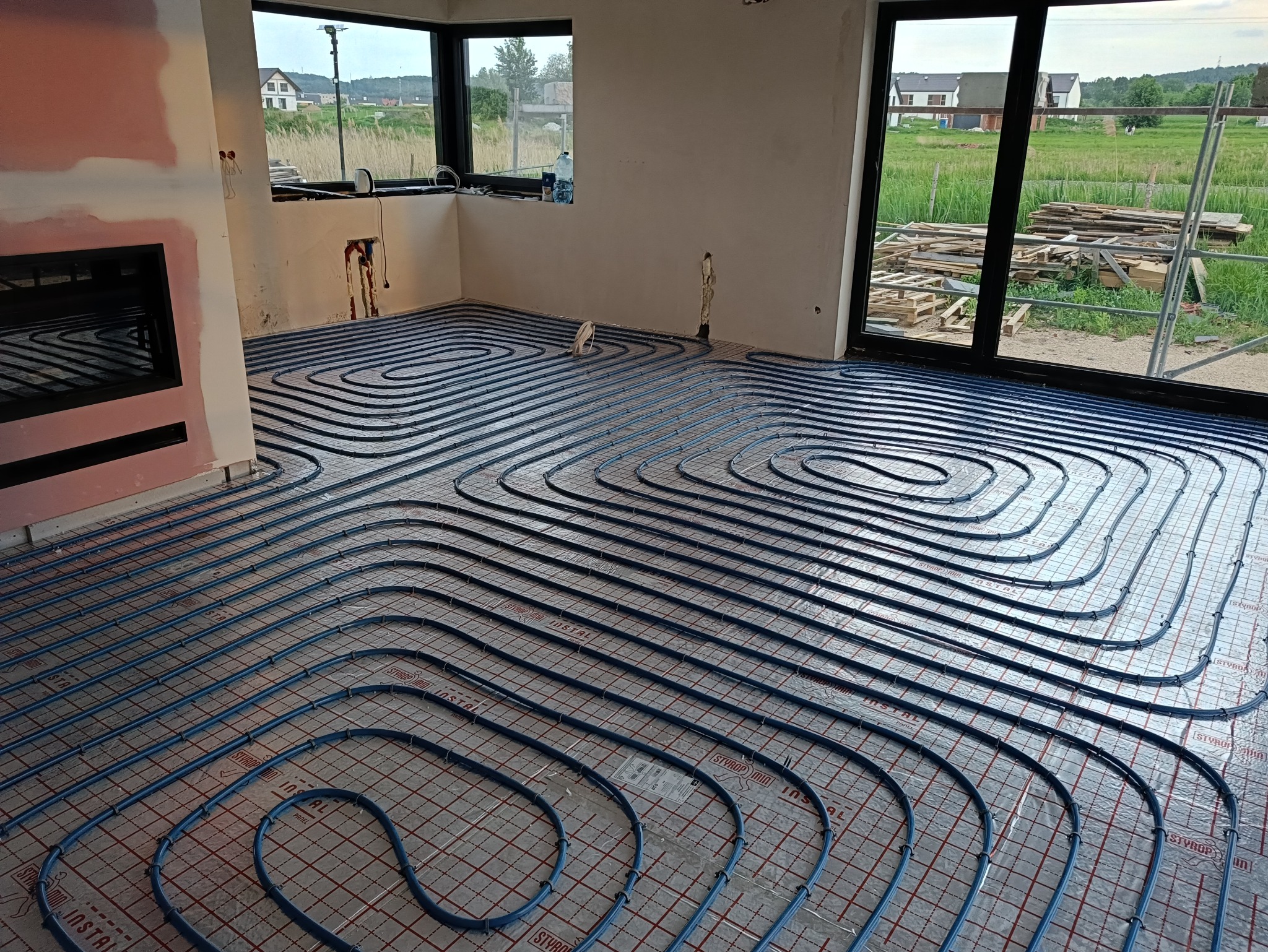 Niebieskie rury ogrzewania podłogowego ułożone spiralnie na srebrnej macie w surowym wnętrzu z dużymi oknami i widokiem na zewnątrz. Etap instalacji przed wylewką.