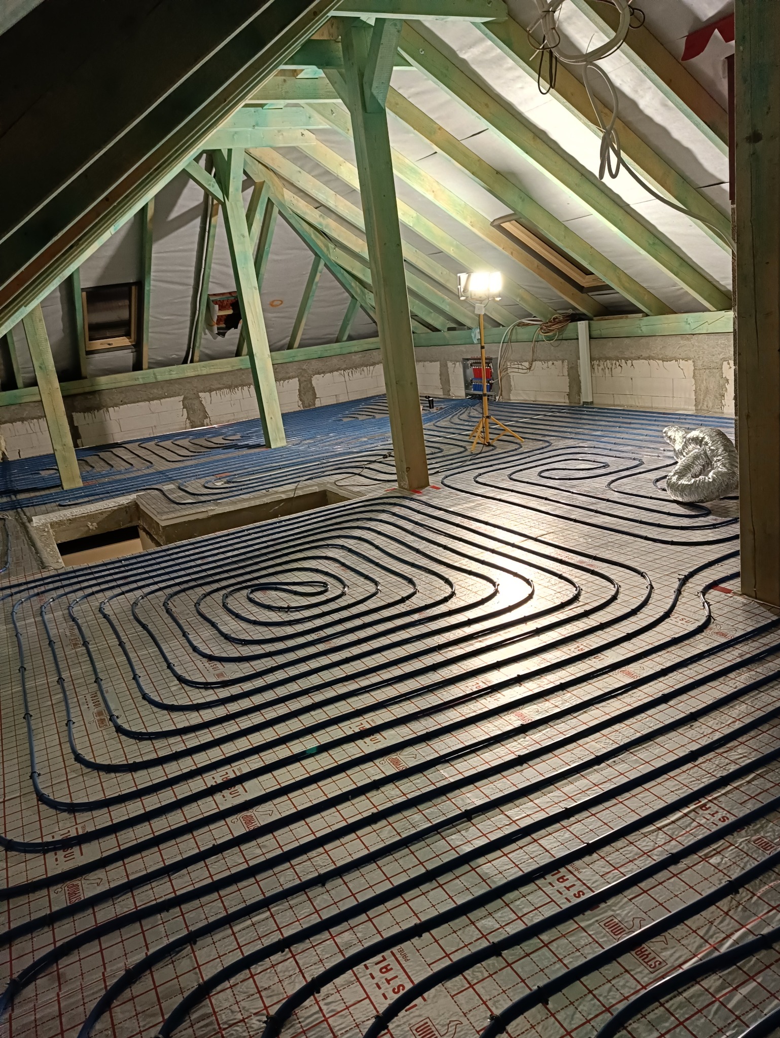 Montaż ogrzewania podłogowego na poddaszu z widoczną konstrukcją dachu i lampą budowlaną. Rury ułożone spiralnie na izolacji z siatką.