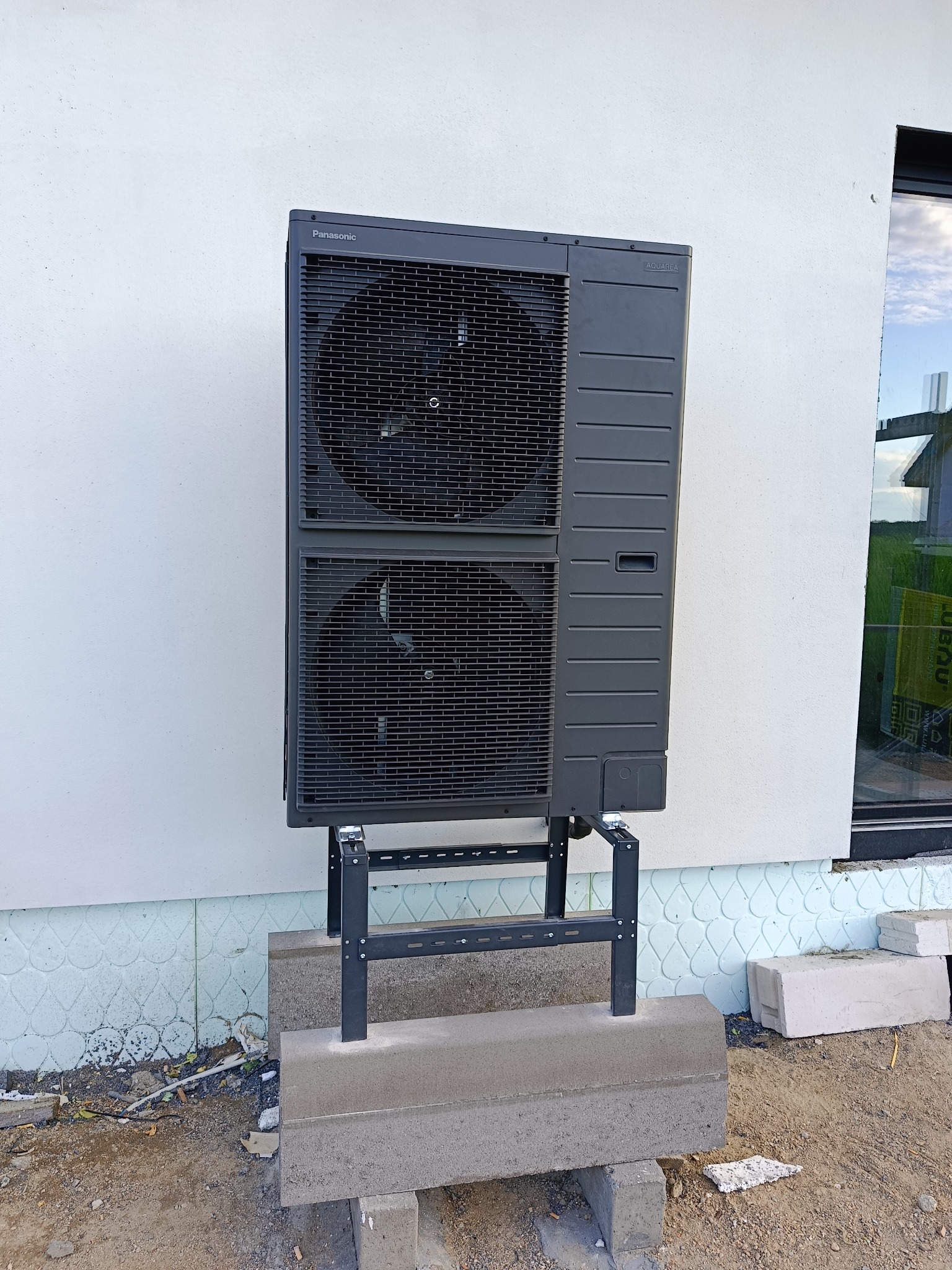 Zewnętrzna jednostka pompy ciepła Panasonic Aquarea zamontowana na wsporniku przy białej ścianie budynku, widoczne elementy instalacji i otoczenie budowy.