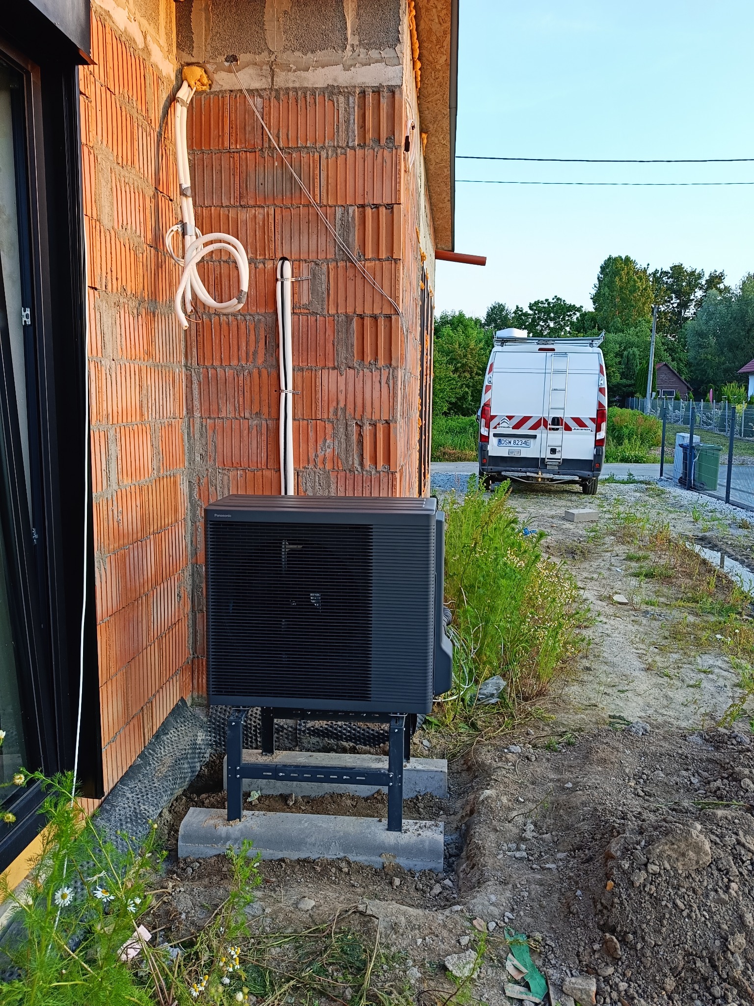 Zewnętrzna jednostka pompy ciepła Panasonic zamontowana przy ścianie budynku z czerwonej cegły, z widocznymi przewodami i samochodem dostawczym w tle.