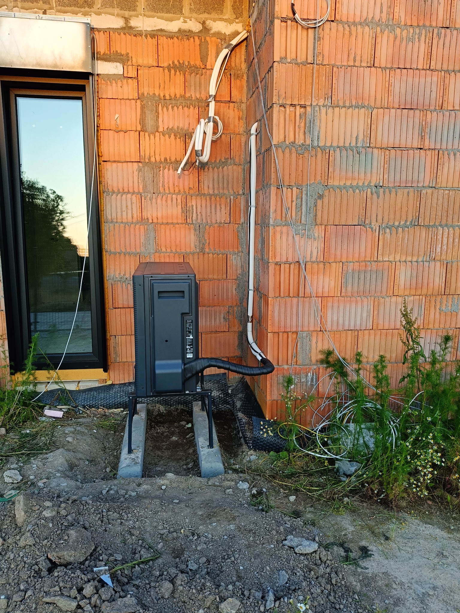 Zewnętrzna jednostka pompy ciepła powietrze-woda, zamontowana na betonowych podstawach przy ścianie budynku z cegły, z widocznymi przewodami i instalacją elektryczną.