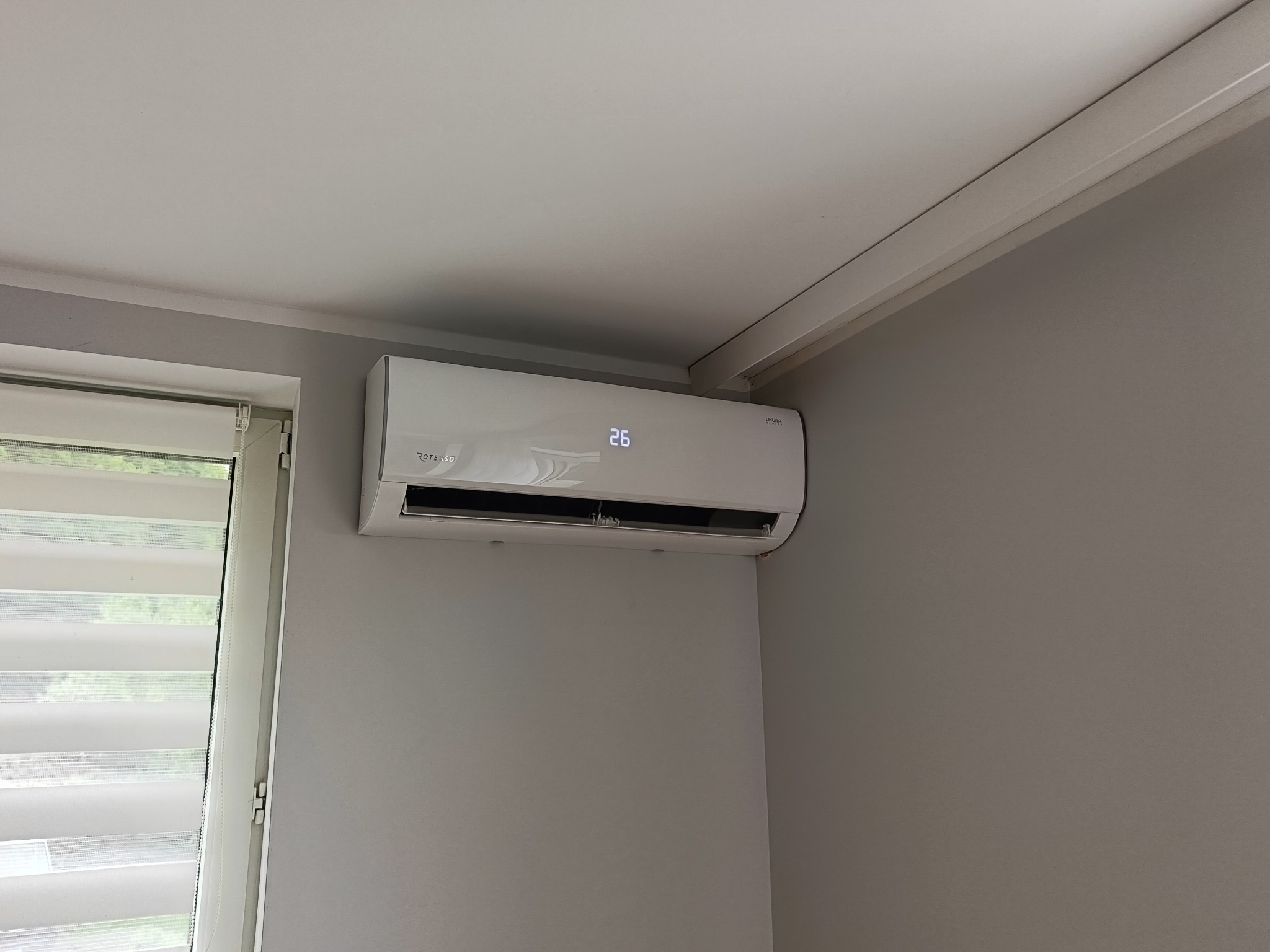 Biały klimatyzator Rotenso z wyświetlaczem 26°C zamontowany na szarej ścianie w pokoju, widok z dołu. Obok okno z żaluzjami dzień-noc.